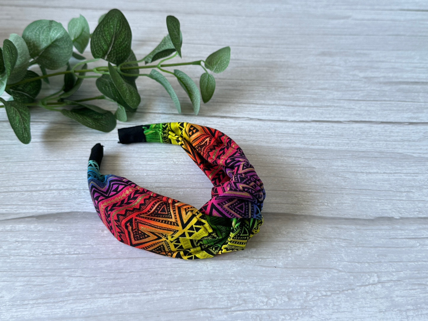 The Cotton Knotted Headband - Rainbow Aztec Print by Crafts by Kate, featuring vibrant geometric designs, rests on a light wooden surface beside green leafy branches.