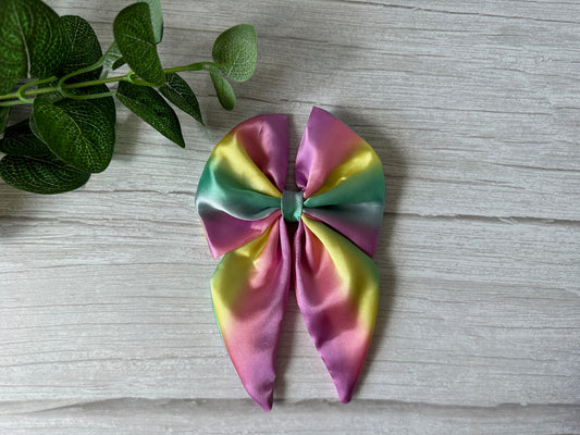 The Silk Sailor Bow Clip - Pastel Rainbow by Crafts by Kate, featuring two tails, is displayed on a light wood surface beside a green leafy plant—adding a luxurious touch and celebrating LGBTIQA+ pride.
