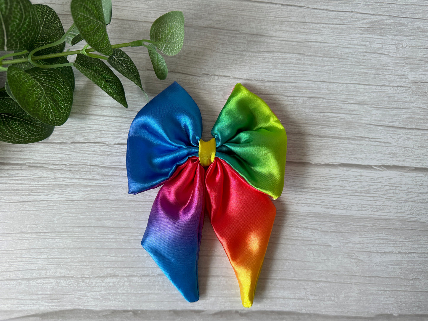 The Silk Sailor Bow Clip - Bright Rainbow by Crafts by Kate, with vibrant rainbow colors, rests on a light wood surface beside green leafy stems—perfect for showing LGBTIQA+ community support.