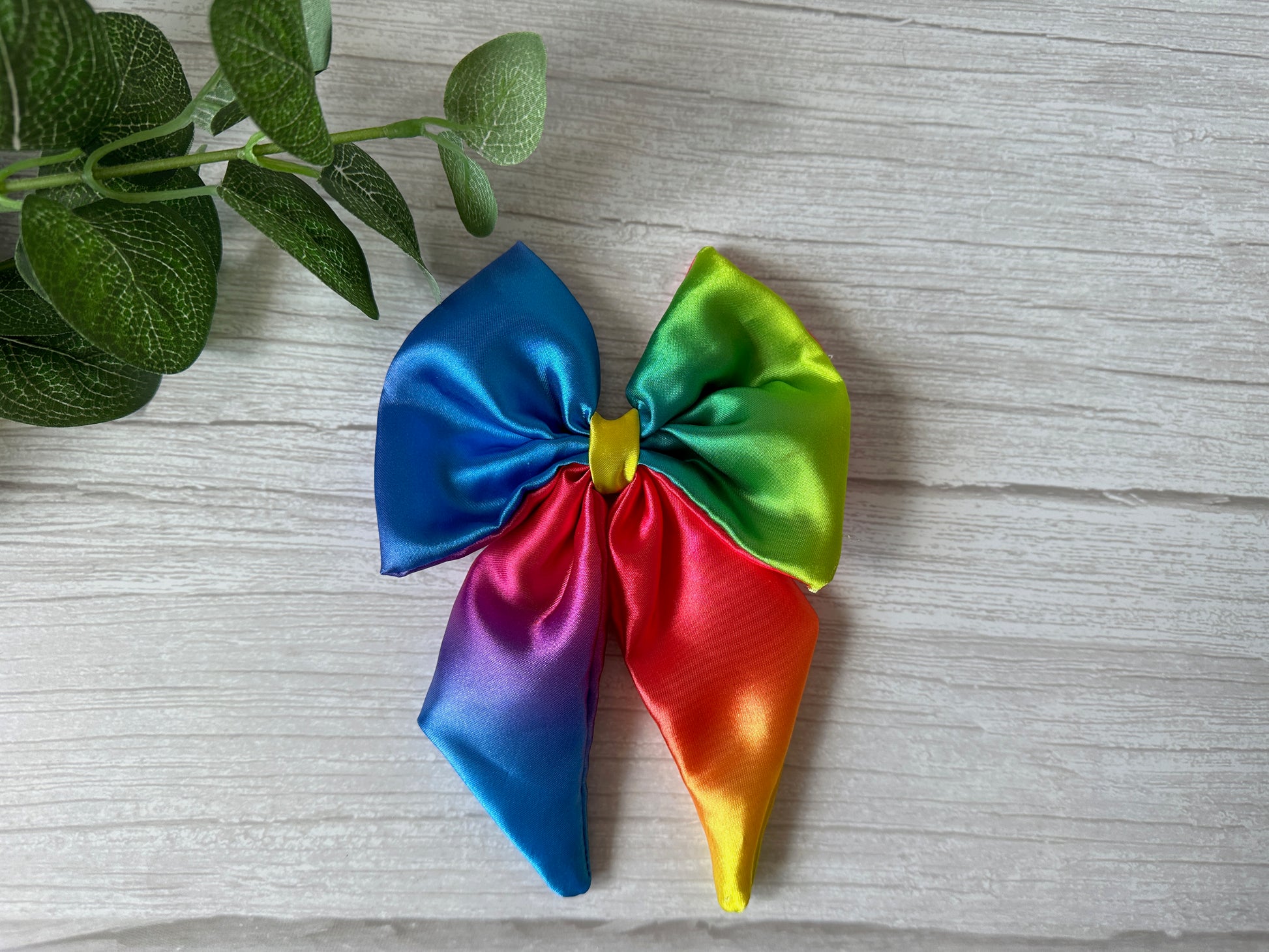 The Silk Sailor Bow Clip - Bright Rainbow by Crafts by Kate, with vibrant rainbow colors, rests on a light wood surface beside green leafy stems—perfect for showing LGBTIQA+ community support.