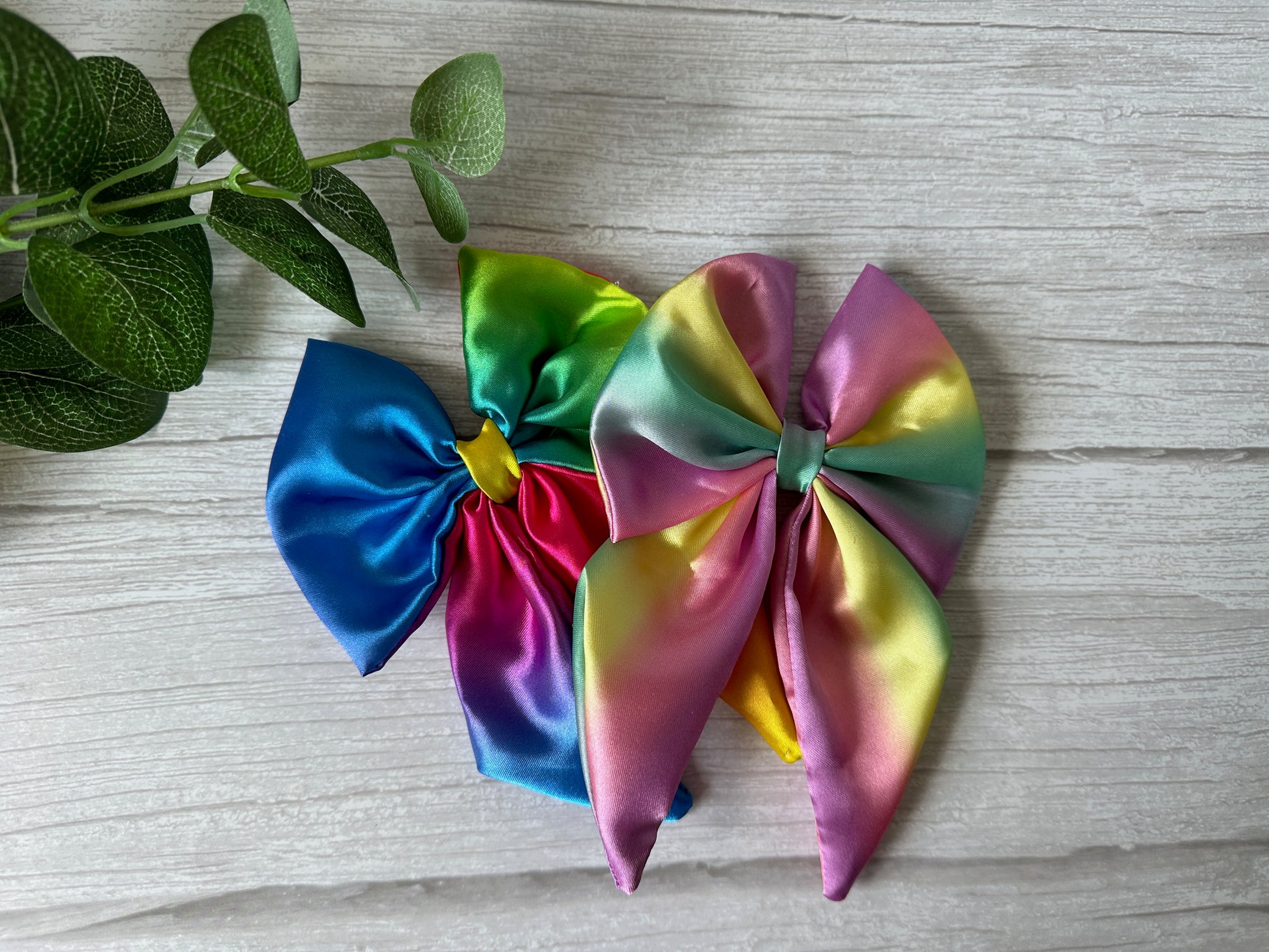 Two vibrant Silk Sailor Bow Clips in a bright rainbow pattern by Crafts by Kate rest on light wood. These colorful accessories subtly show LGBTIQA+ pride. A small green leafy plant appears in the top left corner of the image.
