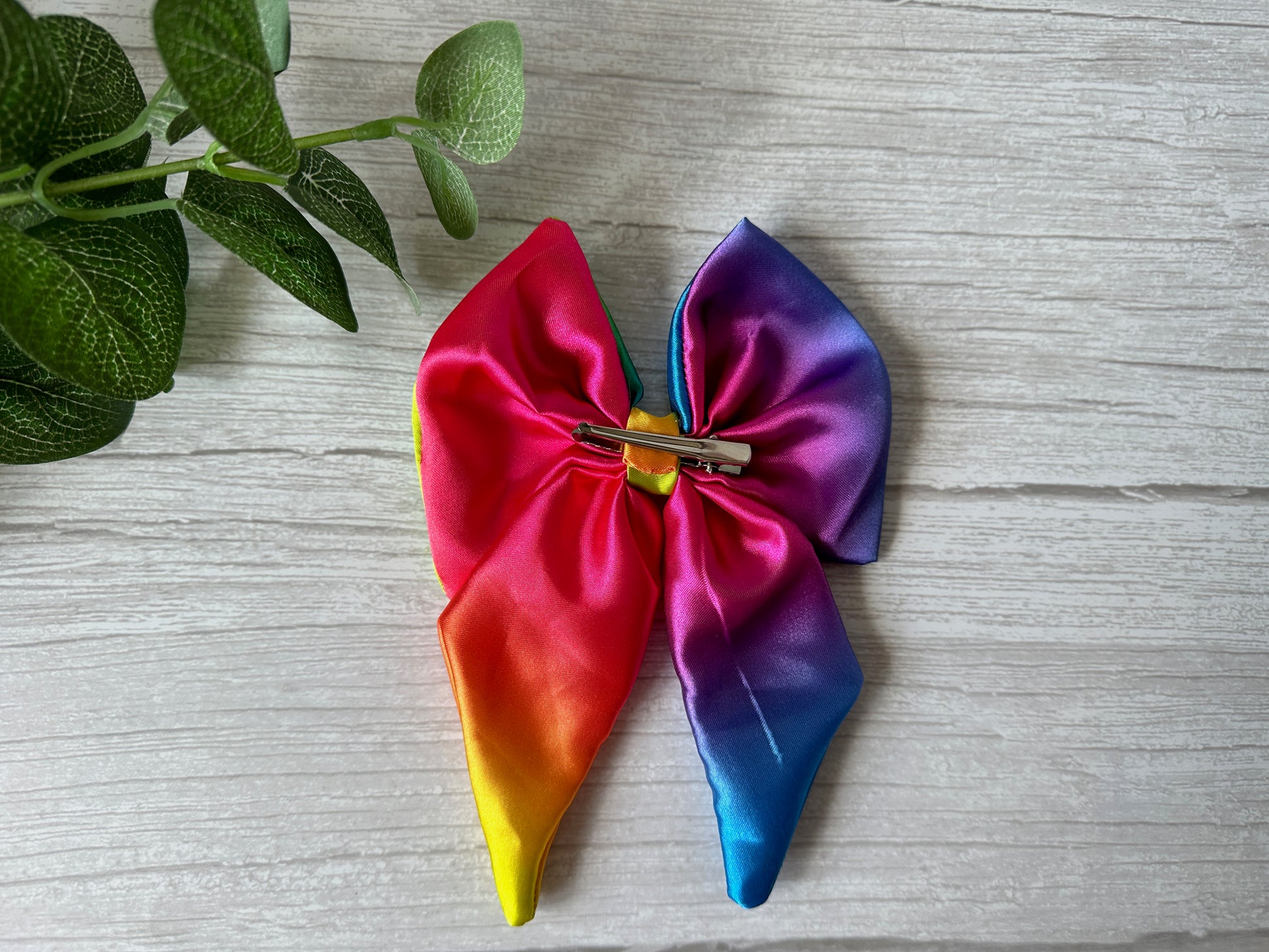 The Silk Sailor Bow Clip - Bright Rainbow by Crafts by Kate, shown on light wood beside a green leafy plant, is a vibrant accessory that elegantly expresses LGBTIQA+ community support.