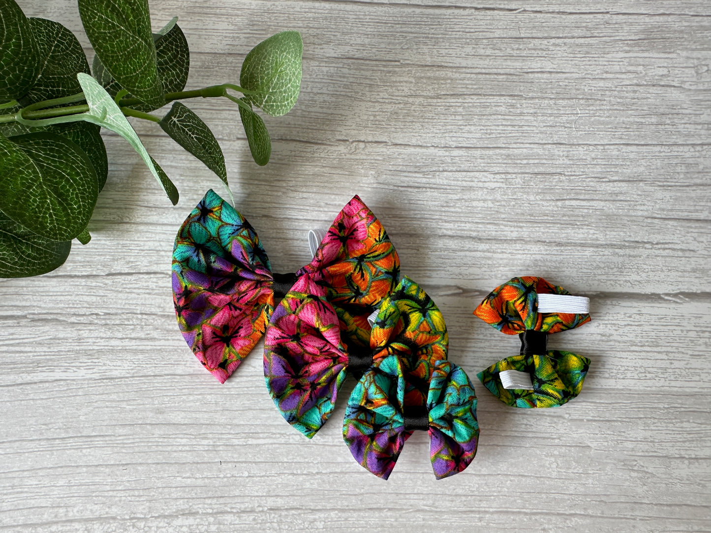 Three colorful hair bows with vibrant floral patterns in purple, green, red, and yellow hues lie on a light wooden surface. A sprig of green leaves decorates the top left corner. One bow is the Cotton Pet Bow with collar attachment - Rainbow Butterfly Print by Crafts by Kate, celebrating the LGBTIQA+ community.