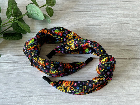 Two black Cotton Knotted Headbands - Rainbow Ginger Cat by Crafts by Kate, made from luxury cotton with colorful floral and botanical patterns, are arranged on a light wood surface with green plant leaves in the top left corner.
