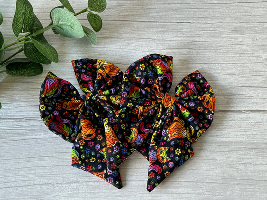 The "Cotton Sailor Bow Clip - Rainbow Ginger Cats" by Crafts by Kate features luxury statement bows with a swirl and floral pattern in orange, pink, green, and yellow against a black background. Displayed on wood with green leaves, they celebrate LGBTIQA+ support.