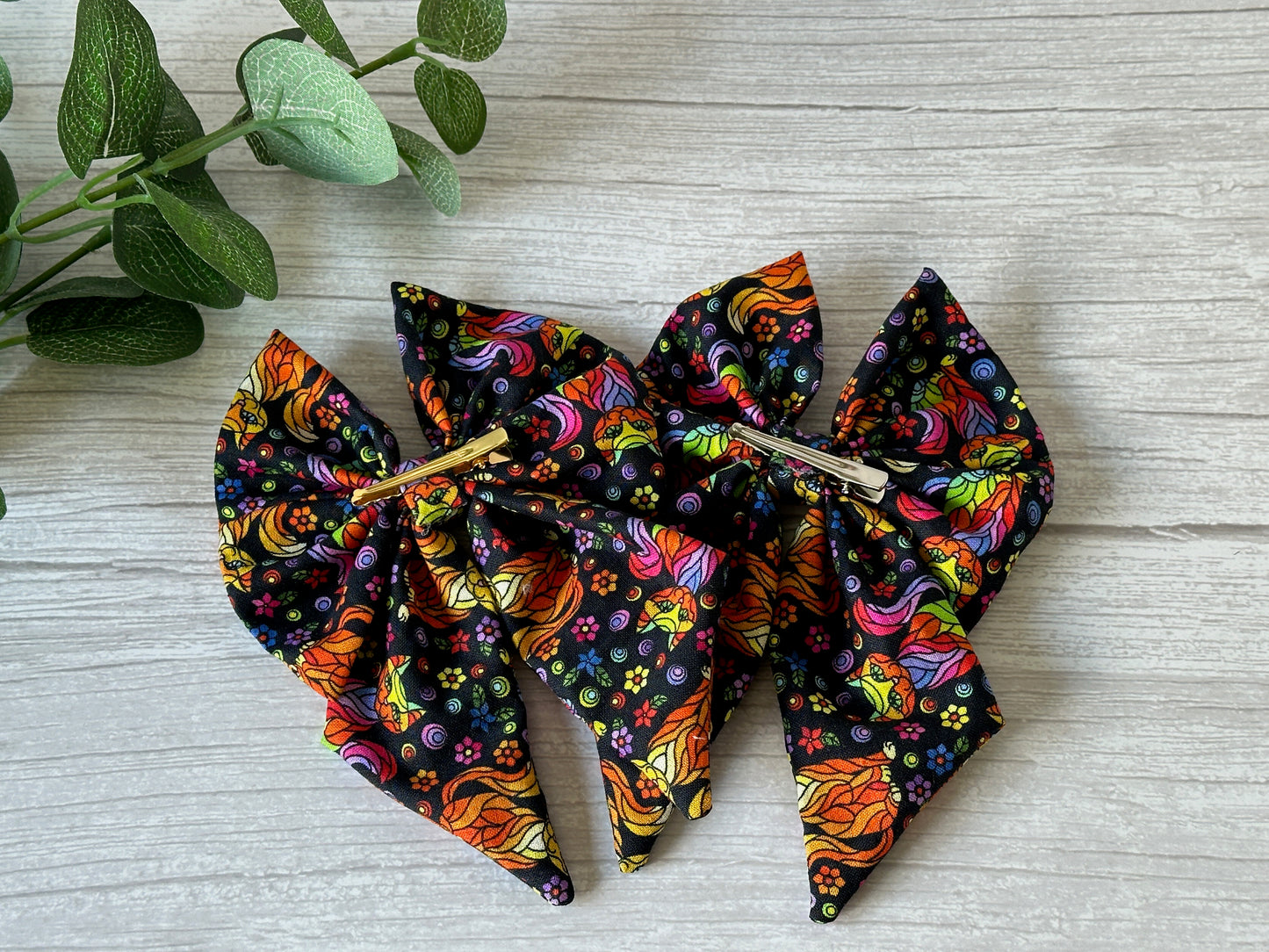 The Cotton Sailor Bow Clip - Rainbow Ginger Cats by Crafts by Kate, adorned with luxury psychedelic swirls and dots, is elegantly set on a light wooden surface accented with green leaves in the top left corner.