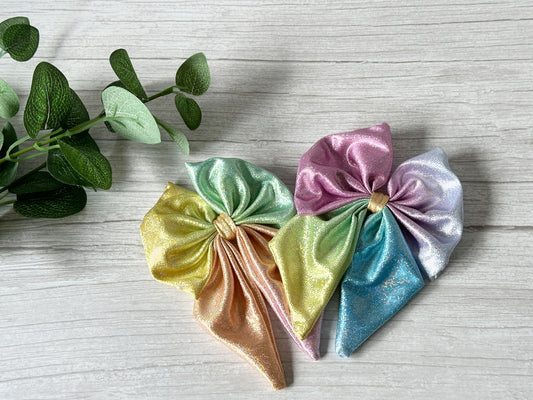 Two vibrant Jersey Foil Hologram Sailor Bow Clips in bright rainbow colors by Crafts by Kate glow on a light wood surface beside a leafy branch—a bold, luxurious statement and subtle nod to LGBTIQA+ support.