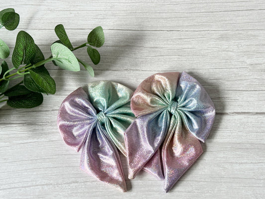 Two large Jersey Foil Hologram Sailor Bow Clips in Mermaid Rainbow by Crafts by Kate, featuring luxury silk style and crocodile clip attachments, rest on a light wood surface beside a green leafy plant.