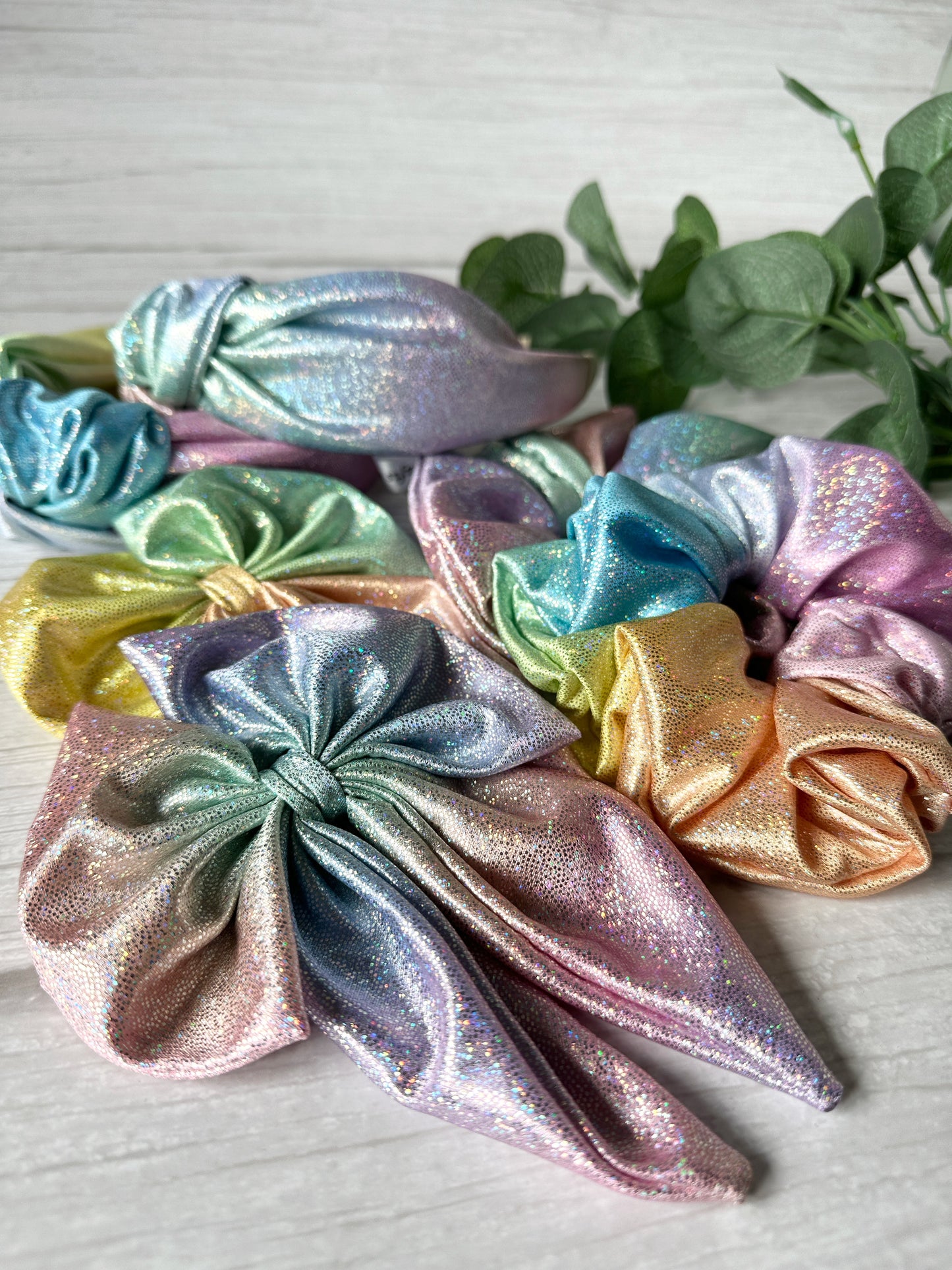 A close-up of Crafts by Kate’s Jersey Foil Hologram Extra Large Scrunchies in Mermaid Rainbow, featuring pastel iridescence and bow tails on a light wood surface, with green leaves adding to the holographic mermaid vibe.