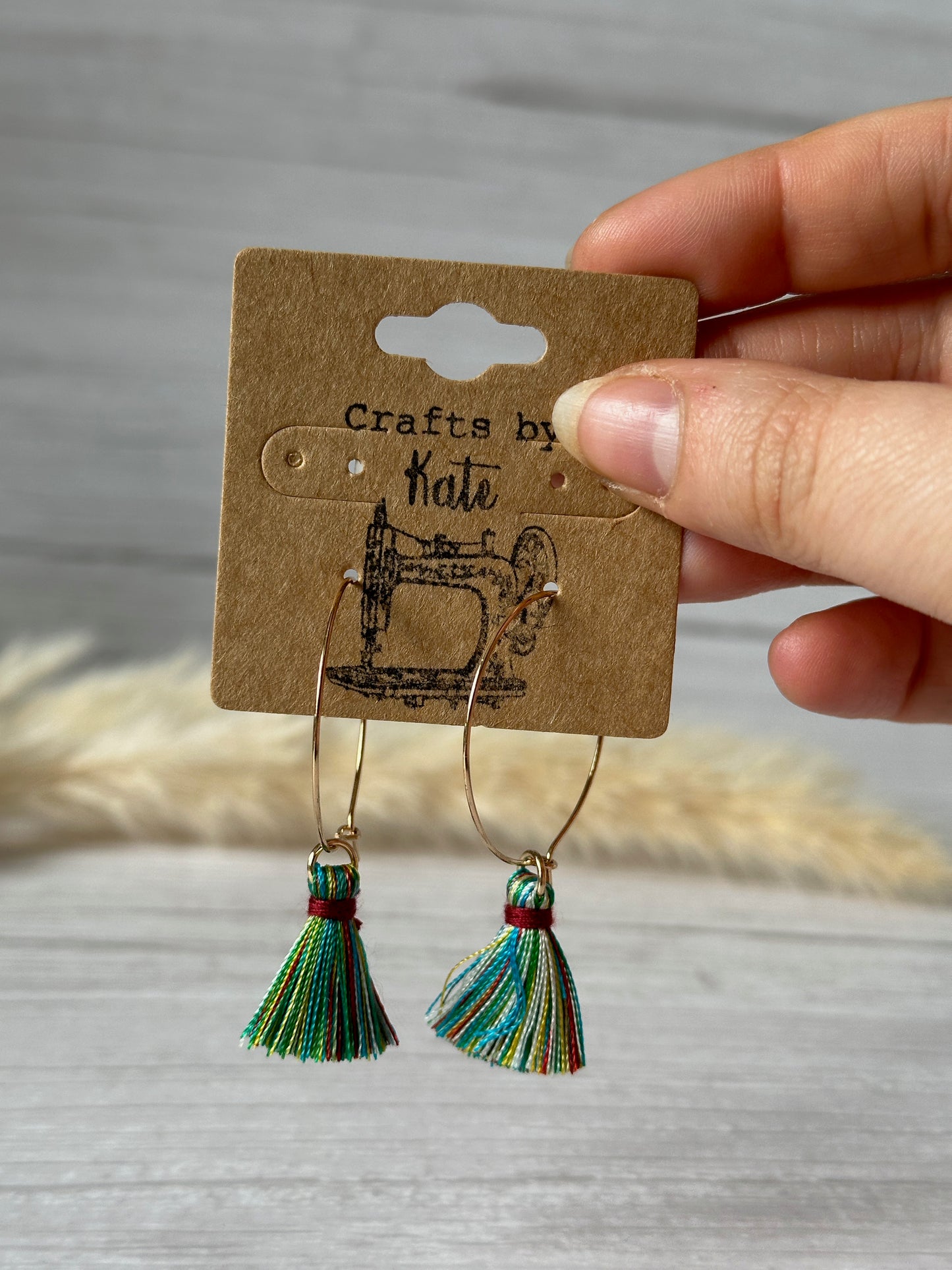 A hand holds vibrant "Crafts by Kate" Handcrafted Rainbow Tassel Gold Plated Hoop Earrings, attached to a brown card with a sewing machine illustration. These eye-catching accessories symbolize spirited LGBTIQA+ community support.