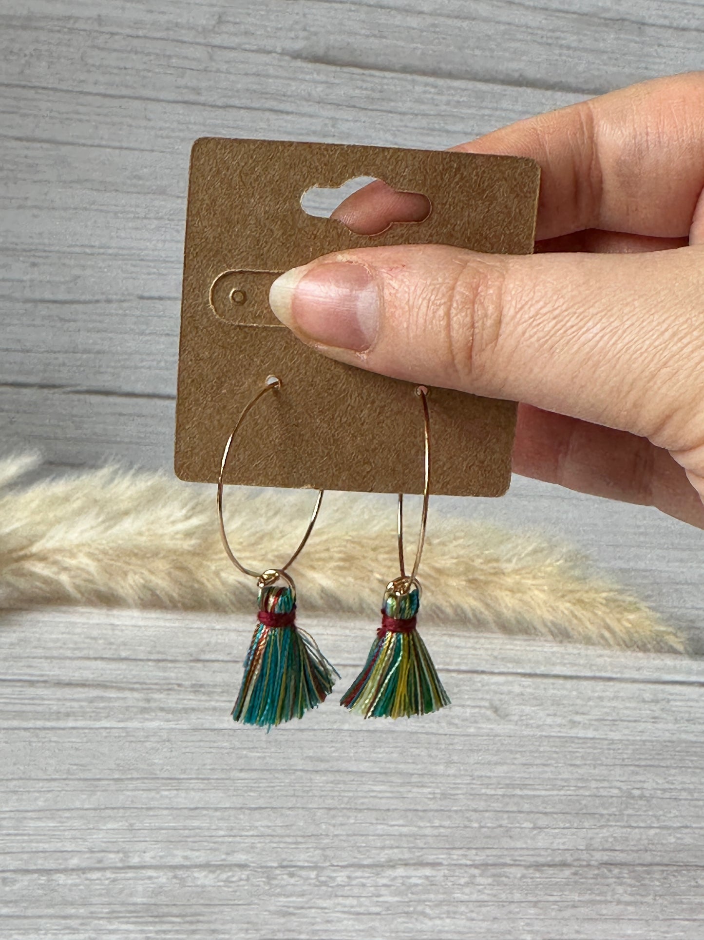 A hand displays a card featuring Crafts by Kate's Handcrafted Rainbow Tassel Gold Plated Hoop Earrings, symbolizing LGBTIQA+ support. The vibrant design is set against a gray wooden backdrop with a fluffy white accent.