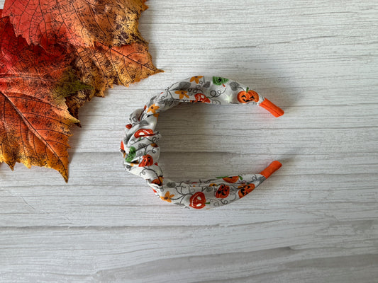 The Cotton Knotted Headband - Pumpkin Lovers by Crafts by Kate, featuring orange tips and a pumpkin pattern, rests on a white wooden surface beside autumn leaves—a cozy accessory for pumpkin enthusiasts.