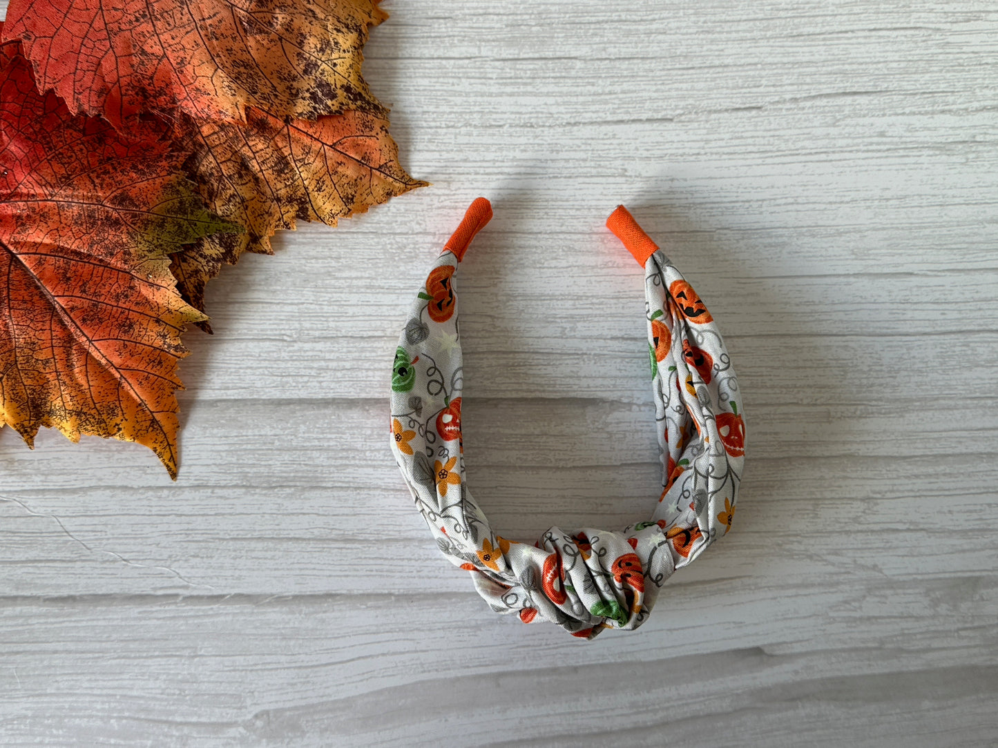 The Cotton Knotted Headband - Pumpkin Lovers by Crafts by Kate, with orange ends and a pumpkin pattern, sits on a light wooden surface next to a large multicolored autumn leaf.