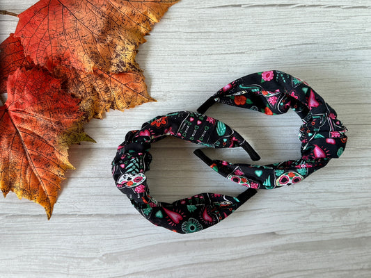Two black Cotton Knotted Headbands - Day of the Dead Cats by Crafts by Kate, featuring colorful cat skulls and floral designs, rest on a light wooden surface with orange autumn leaves nearby.