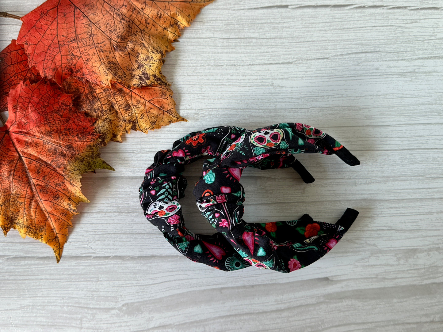 The Crafts by Kate Cotton Knotted Headband - Day of the Dead Cats, featuring playful cat skulls and floral patterns, is displayed on a light wooden surface beside two orange and red autumn leaves.