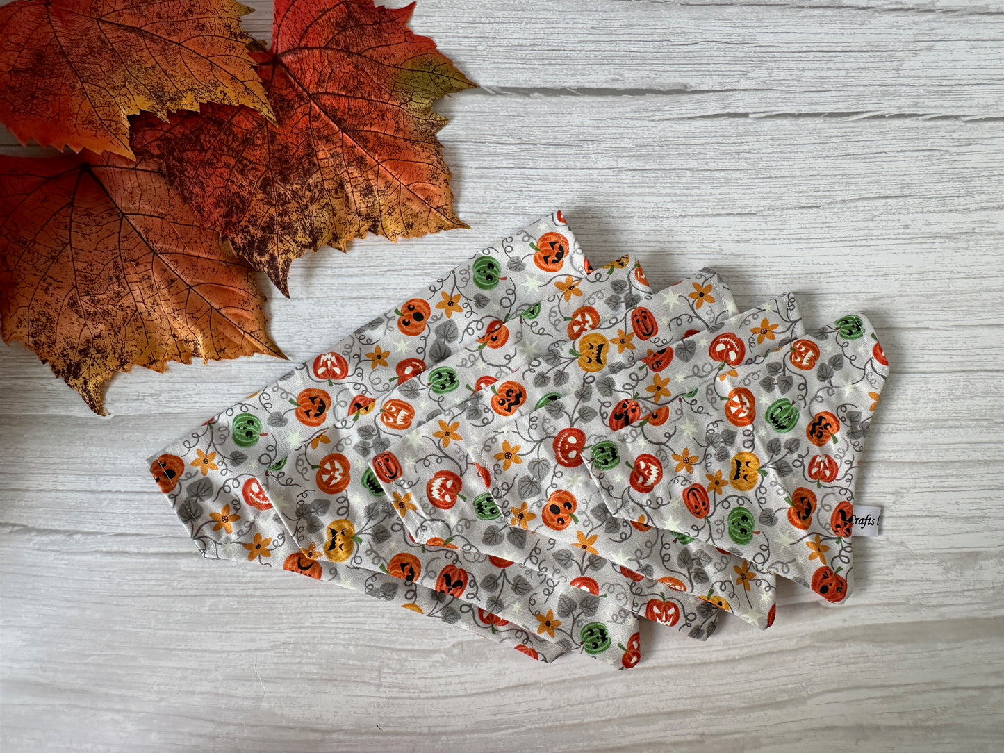 A Cotton Pet Bandana with collar attachment—Glow in the Dark Pumpkins by Crafts by Kate rests on a white wooden surface beside several artificial autumn leaves.