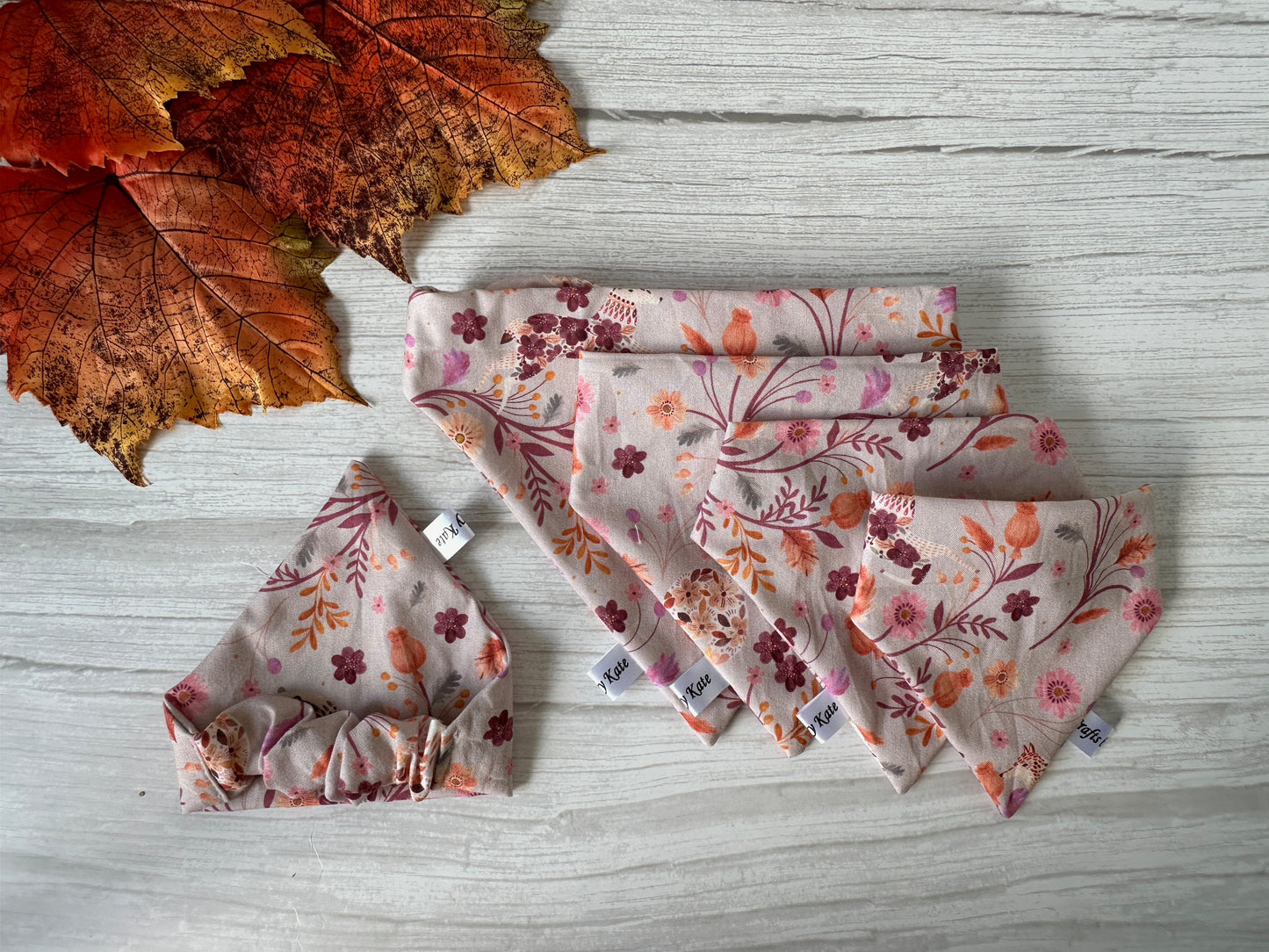 Neatly folded on a wooden surface, the Cotton Pet Scrunchie Bandana - Woodland Floral by Crafts by Kate features an enchanting pink and red floral design. This seasonal accessory evokes a warm, cozy charm reminiscent of fall, complemented by autumn leaves in the top left corner.