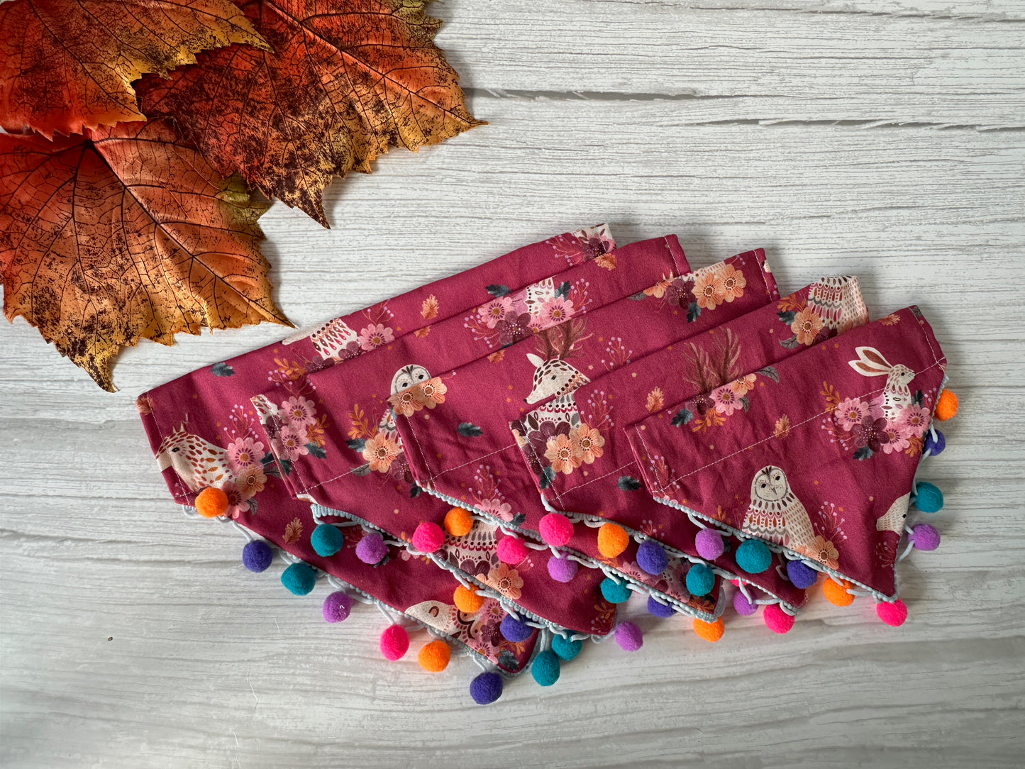 A Cotton Pet Bandana with collar attachment by Crafts by Kate, featuring woodland animals and colorful pom poms, is displayed on a light wood surface next to artificial autumn leaves.