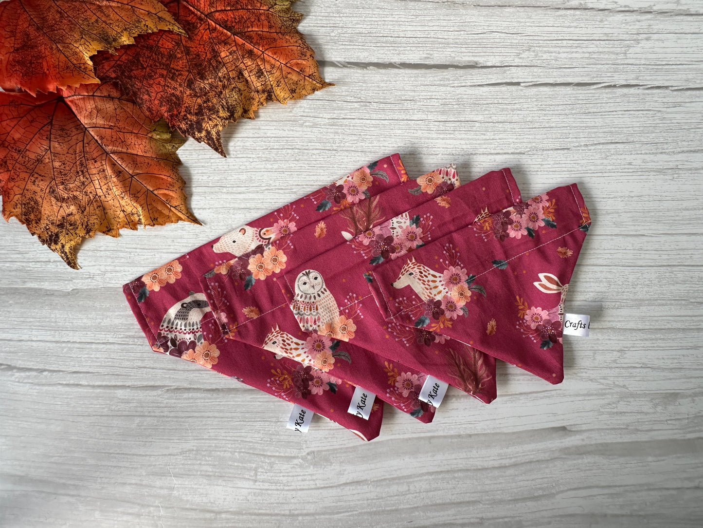 Three Crafts by Kate Cotton Pet Bandana with collar attachment – Woodland Animals designs, featuring maroon fabric with floral and owl patterns, are displayed on a white wooden surface. Each bandana has a small white tag; faux autumn leaves accent the top left corner.