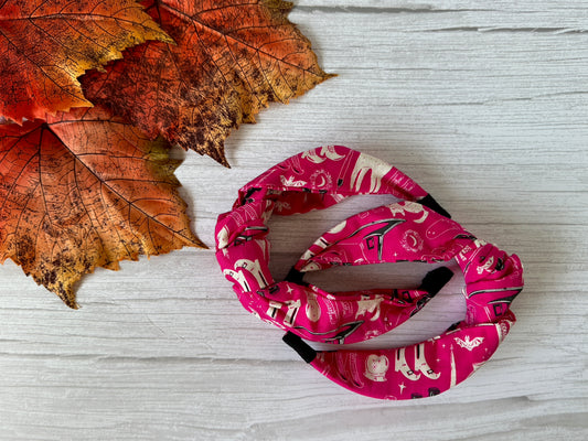 The Crafts by Kate Cotton Knotted Headband - Pink Witches, featuring white and red printed designs, rests on a white wooden surface beside orange and red autumn leaves.
