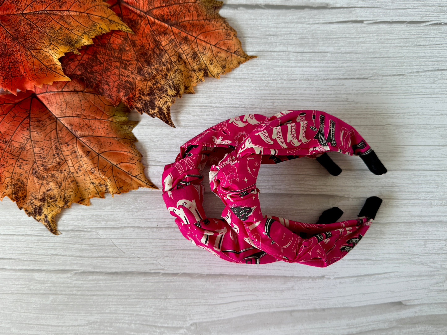 A pair of Crafts by Kate Cotton Knotted Headband - Pink Witches rests on a white wooden surface beside orange autumn leaves.