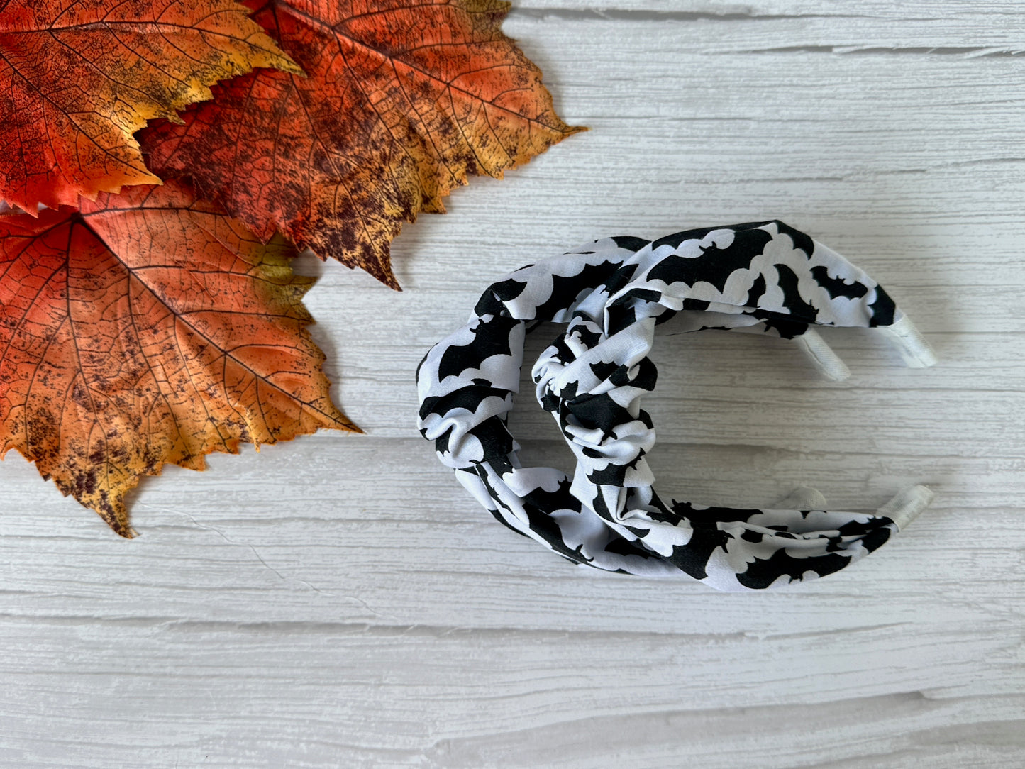 Two Cotton Knotted Headbands - Bats by Crafts by Kate are displayed on a white wooden surface beside autumn-colored artificial maple leaves.