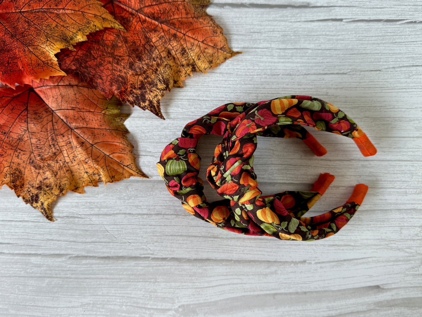 Two headbands from Crafts by Kate, including the Cotton Knotted Headband - Pumpkin Lovers with an autumn-inspired print, rest on a white wooden surface beside orange and red fall leaves.