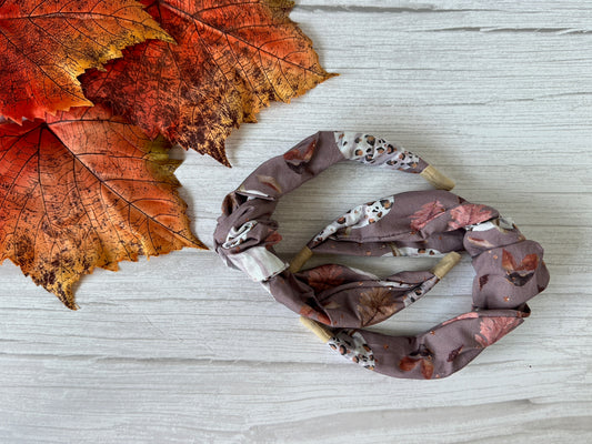 The Cotton Knotted Headband - Leopard Print Pumpkins by Crafts by Kate rests on a light wooden surface beside vibrant orange and red artificial maple leaves, adding handcrafted charm to your fall look.