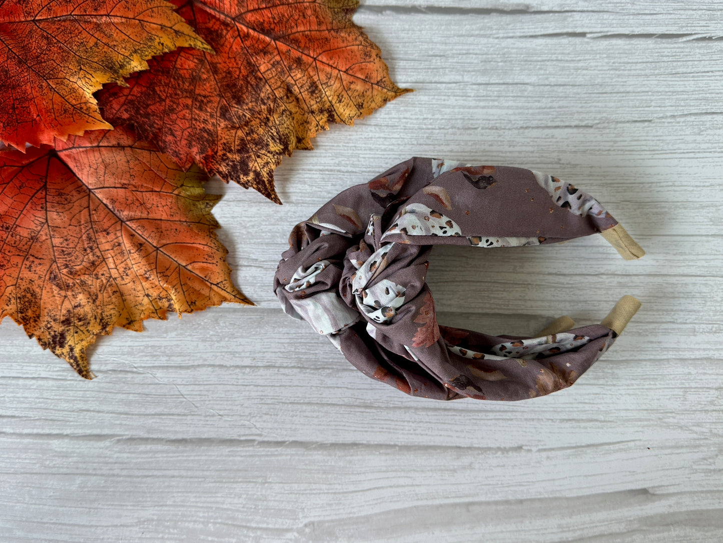 The Cotton Knotted Headband - Leopard Print Pumpkins by Crafts by Kate sits on a white wooden surface, accented by artificial autumn leaves in orange and red.