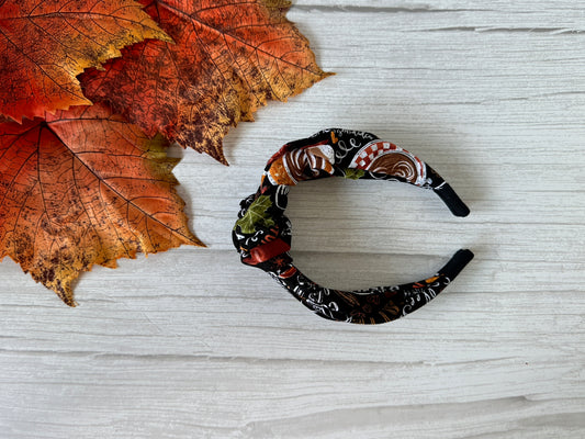 The Crafts by Kate Cotton Knotted Headband - Pumpkin Spice, featuring an autumn-inspired design, is displayed on a white wooden surface next to orange and red artificial fall leaves.