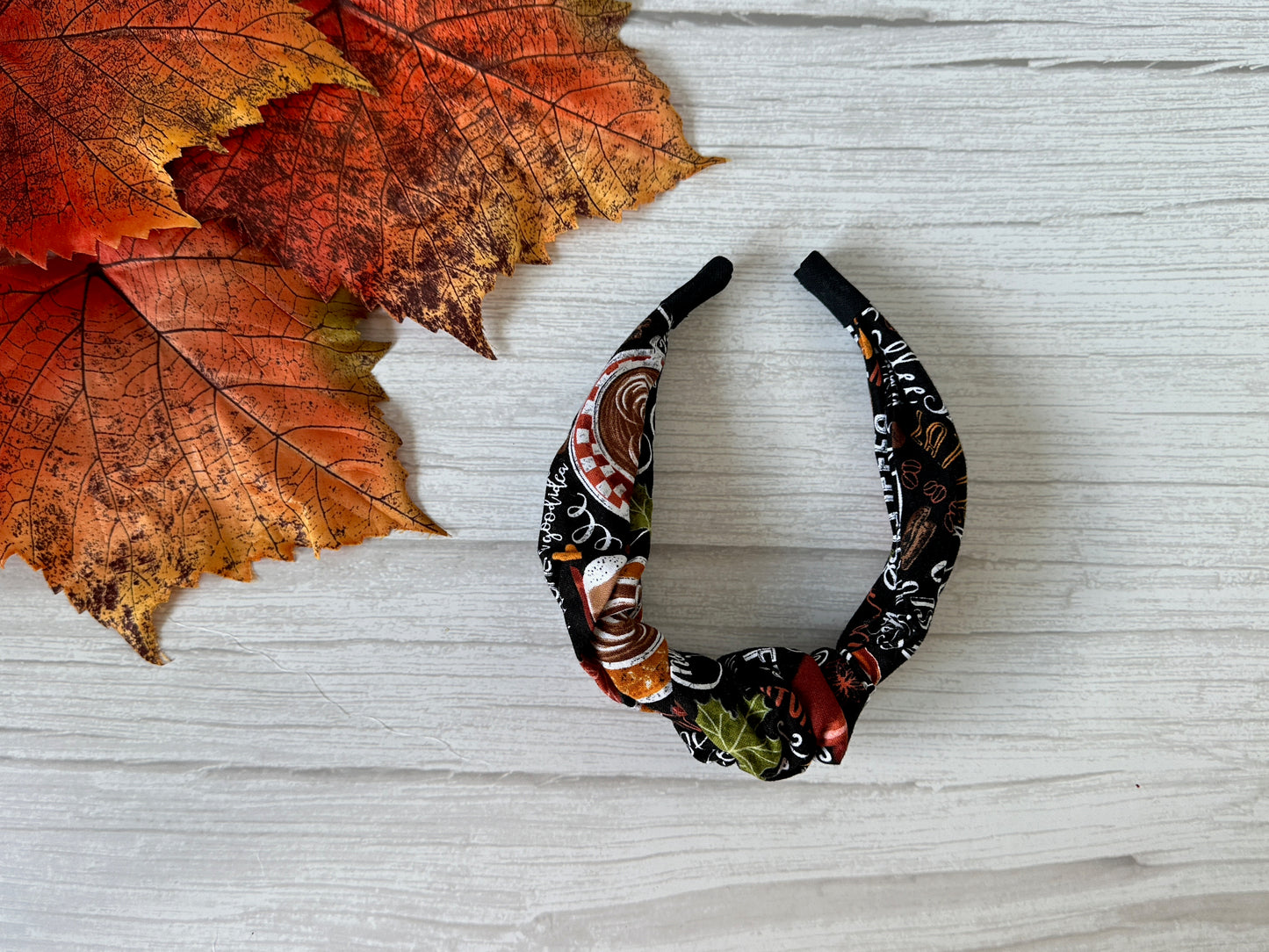 The Crafts by Kate Cotton Knotted Headband - Pumpkin Spice, featuring autumn patterns, rests on a white wooden surface beside orange and red fall leaves.