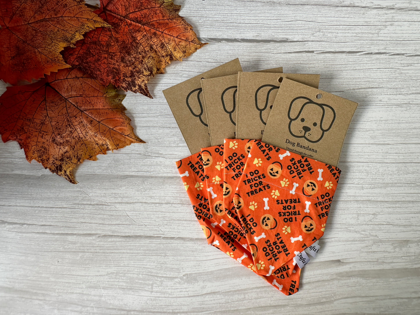 Four orange cotton pet bandanas by Crafts by Kate, featuring collar attachments and "I do tricks for treats" text, are displayed on a wooden surface with autumn leaves. Each handcrafted bandana includes a dog-illustrated "Dog Bandana" tag.