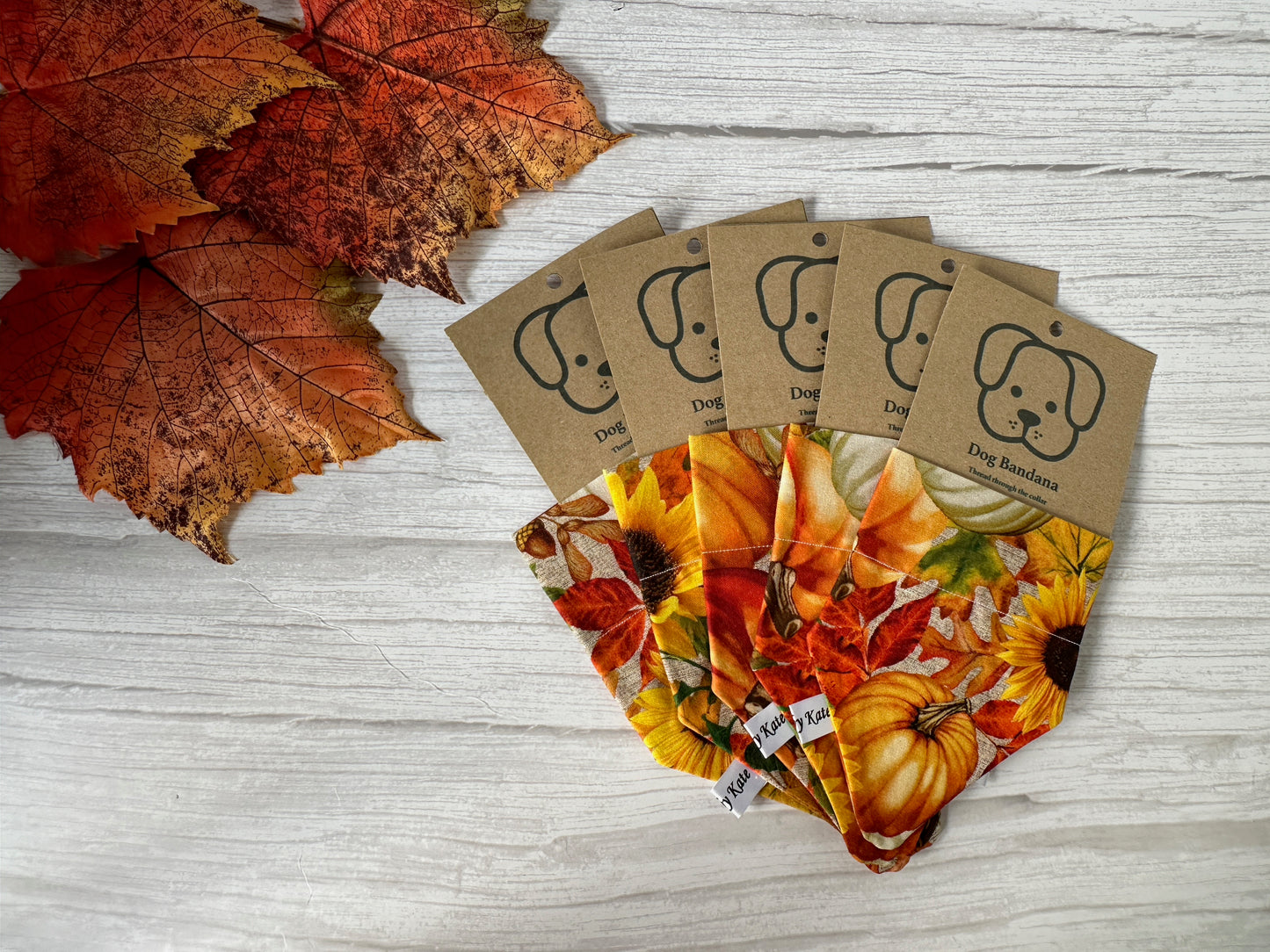 Crafts by Kate's Cotton Pet Bandana with collar attachment - Sunflower Pumpkins showcases vibrant sunflower and leaf designs crafted from autumn-themed fabric. Each cotton accessory features a tag displaying a cartoon dog face, and the display is complemented by a cluster of colorful fall leaves on a rustic wooden surface.