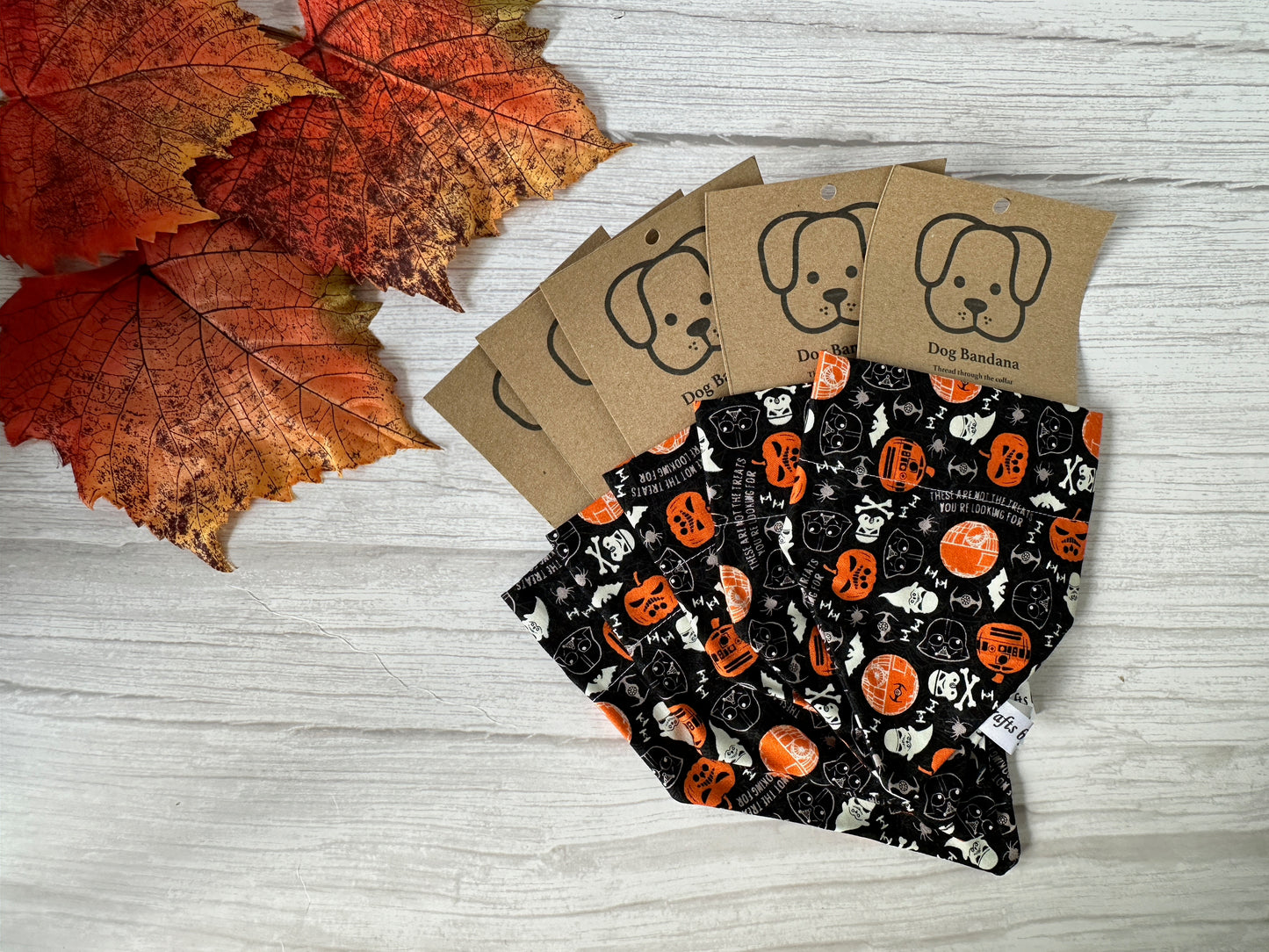Five black "Cotton Pet Bandana with collar attachment - Star Wars The Dark Side" by Crafts by Kate, each on a dog-illustrated backing, are displayed on a white wooden surface beside autumn leaves.