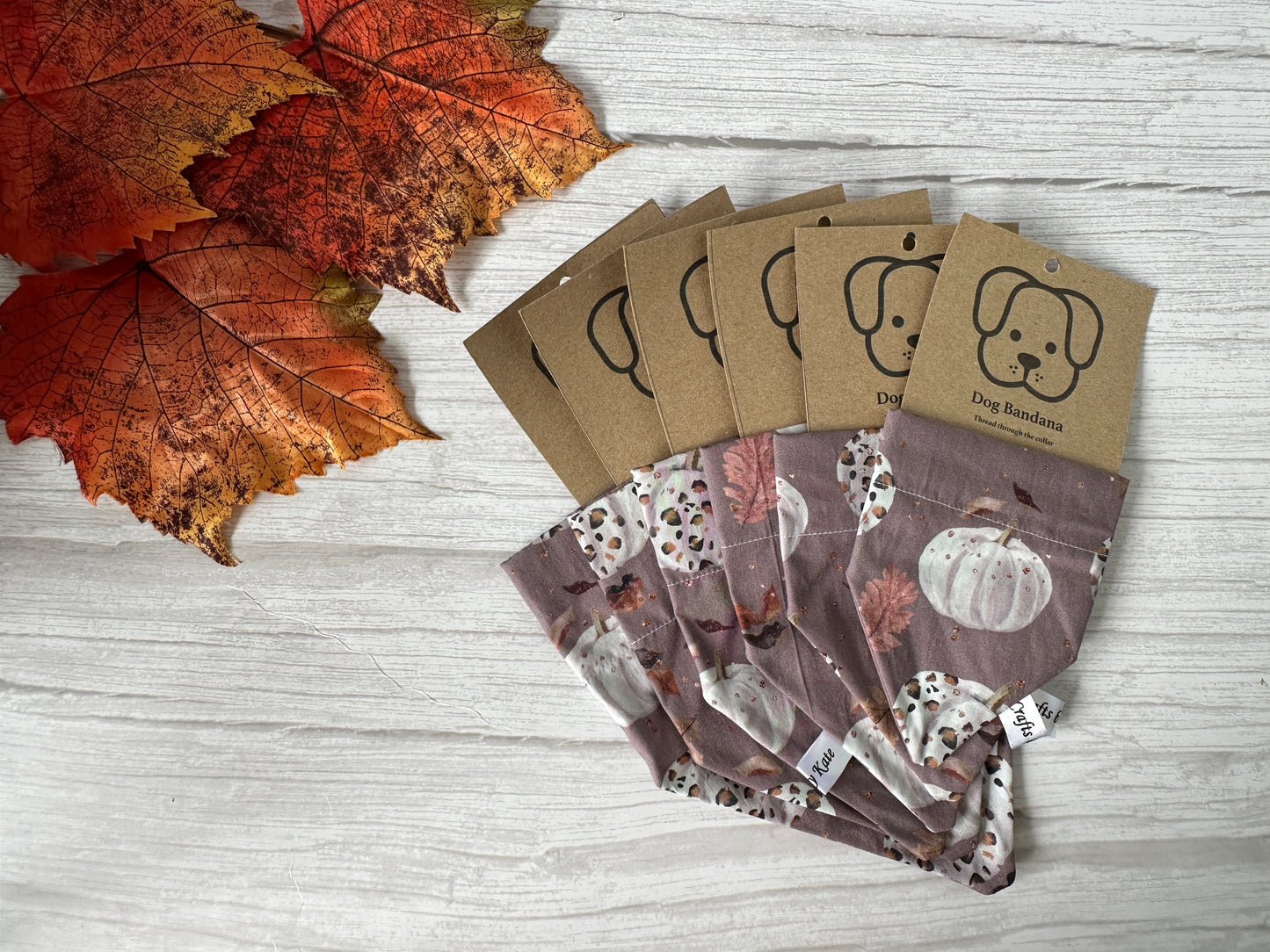 Five Crafts by Kate Cotton Pet Bandanas with collar attachment, featuring leopard print pumpkins, are displayed on a white wooden surface beside three artificial fall leaves.