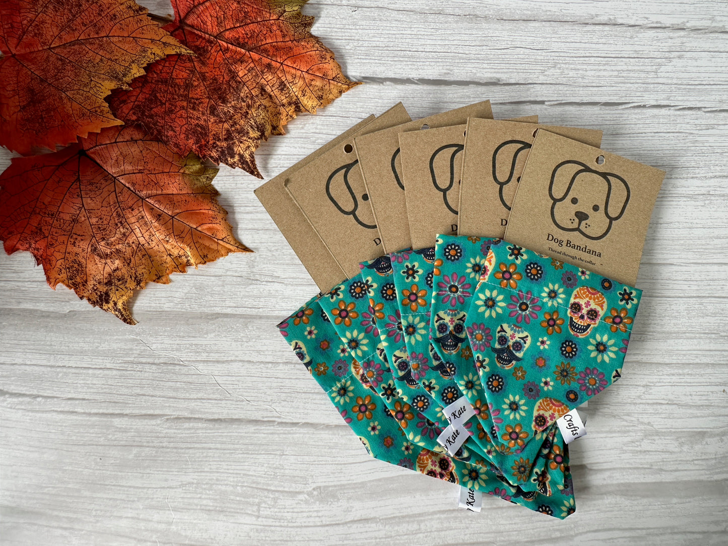 Four Crafts by Kate "Cotton Pet Bandana with collar attachment - Day of the Dead" bandanas with floral and Day of the Dead designs, each with a dog-tag label, are displayed near autumn leaves on a light wooden surface.