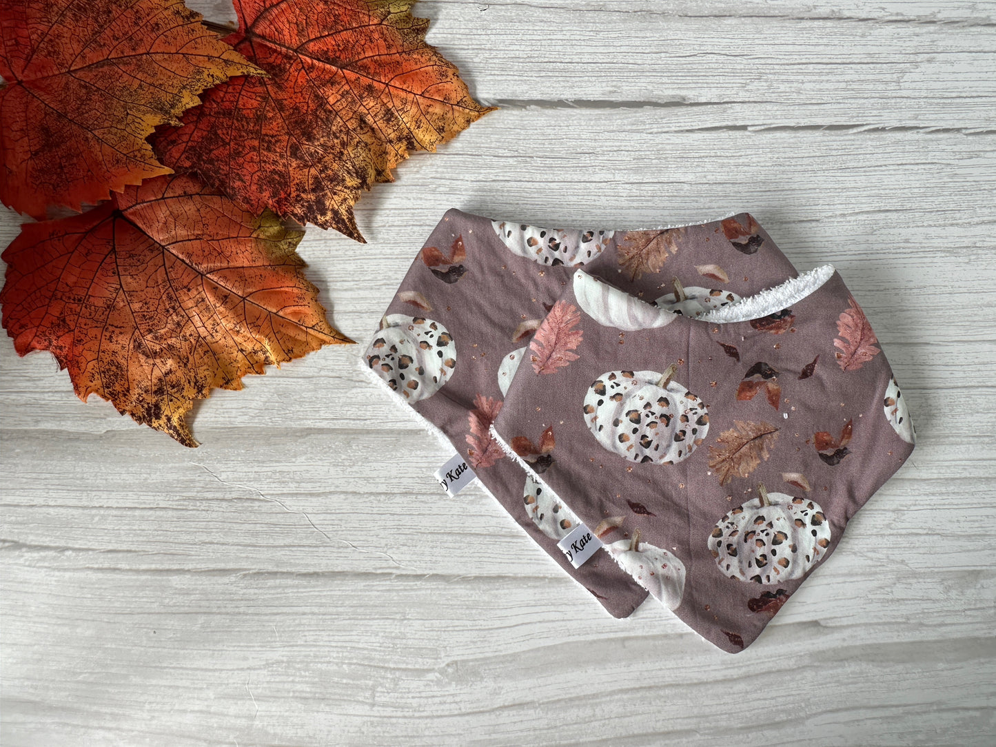 The Cotton & Terry Towelling Baby Bandana Bib - Leopard Print Ghosts by Crafts by Kate is displayed on a light wood surface beside several orange-red fall leaves.