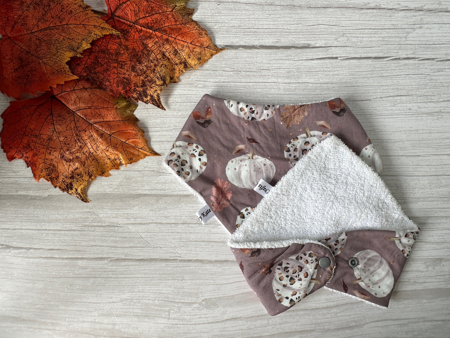 The Cotton & Terry Towelling Baby Bandana Bib - Leopard Print Ghosts by Crafts by Kate features a spooky design, with soft cotton and absorbent terry backing, styled on a white wood surface next to orange autumn leaves.