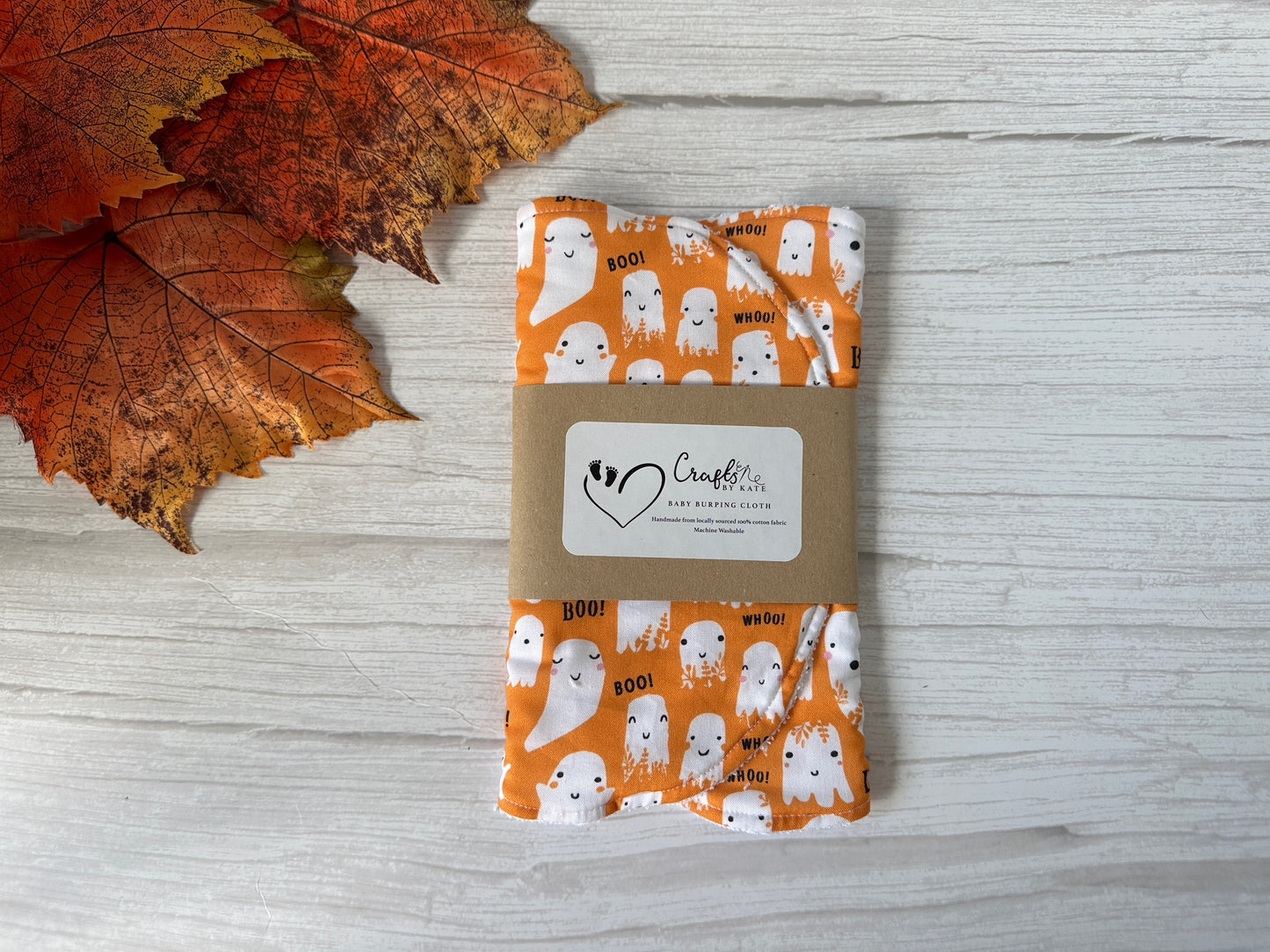 The Cotton & Terry Towelling Baby Burping Cloth - Baby Ghosts by Crafts by Kate, featuring playful ghosts and "BOO!" design, is folded with a brown label and set on a white wooden surface next to orange autumn leaves.