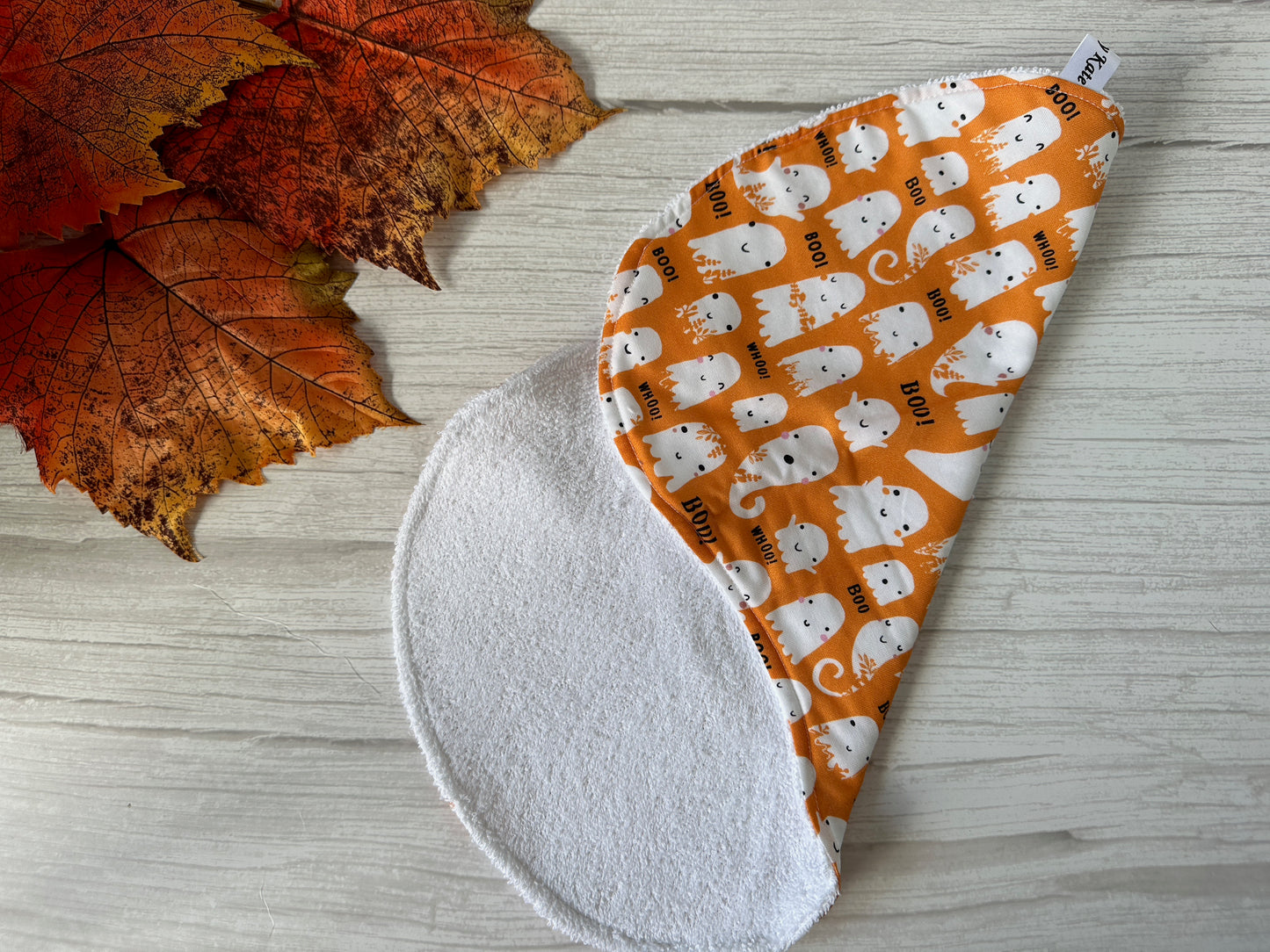 The Cotton & Terry Towelling Baby Burping Cloth - Baby Ghosts by Crafts by Kate, featuring soft terry on one side and a playful ghost print on the other, is shown on a light wood surface with autumn leaves nearby.