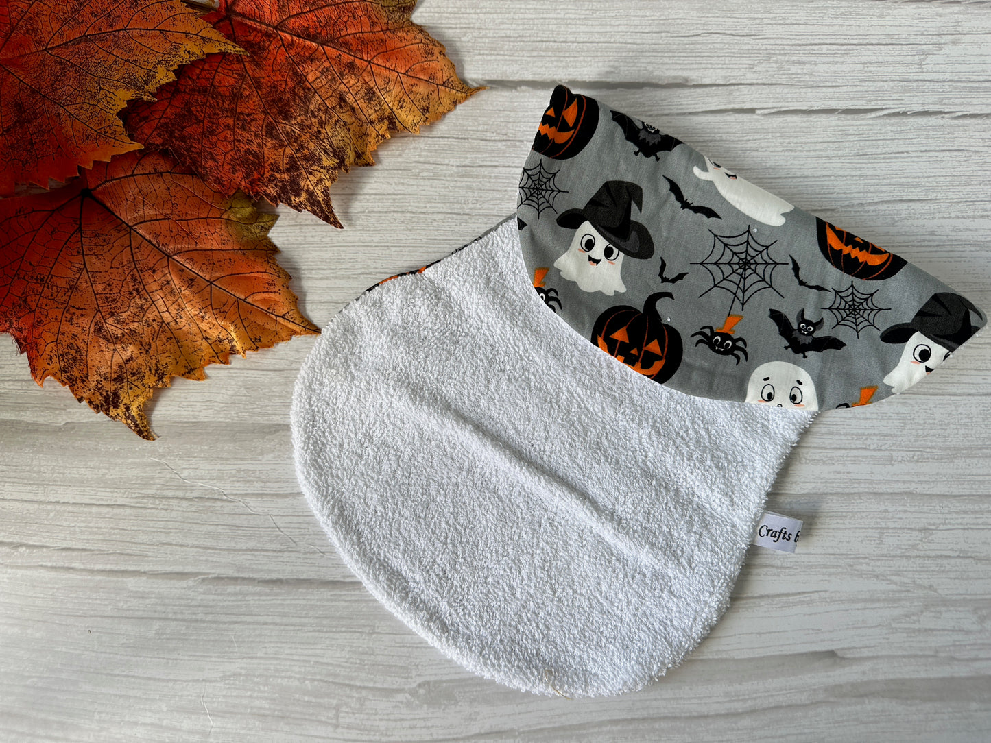 The Cotton & Terry Towelling Baby Burping Cloth - Grey Halloween by Crafts by Kate features ghosts, pumpkins, bats, and spiderwebs, shown with autumn leaves on a white wooden surface.