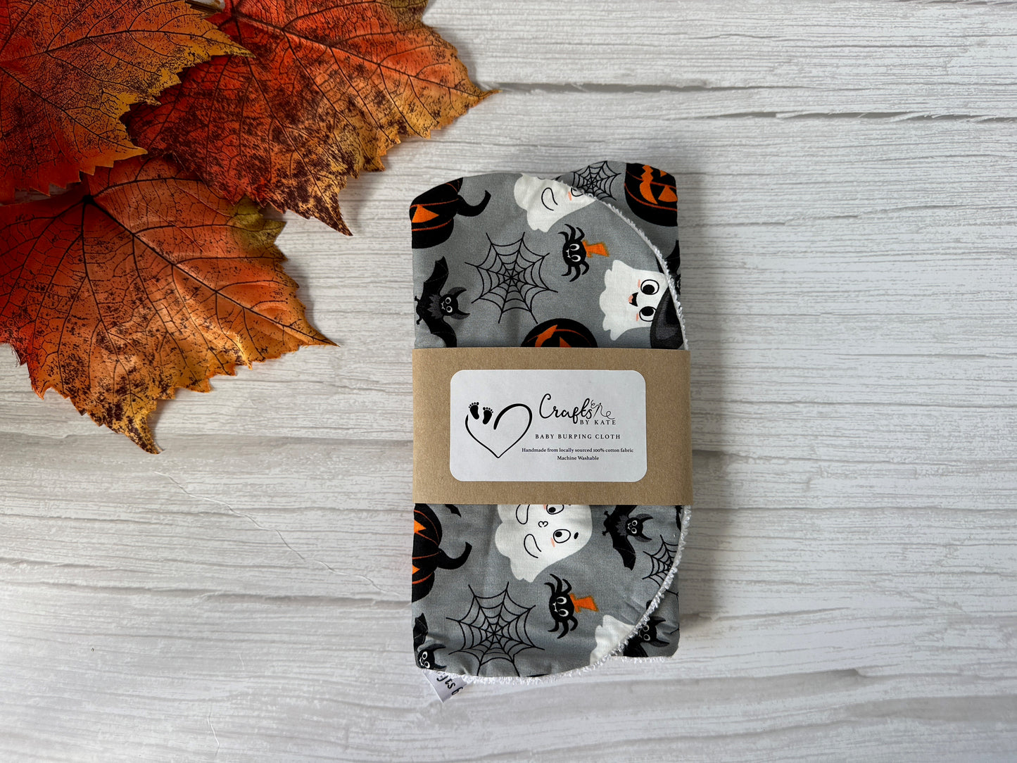 A bundle of Crafts by Kate Cotton & Terry Towelling Baby Burping Cloths in a grey Halloween print—featuring ghosts, bats, pumpkins, and spiderwebs—displayed with autumn leaves and wrapped in a branded label on a white wooden surface.