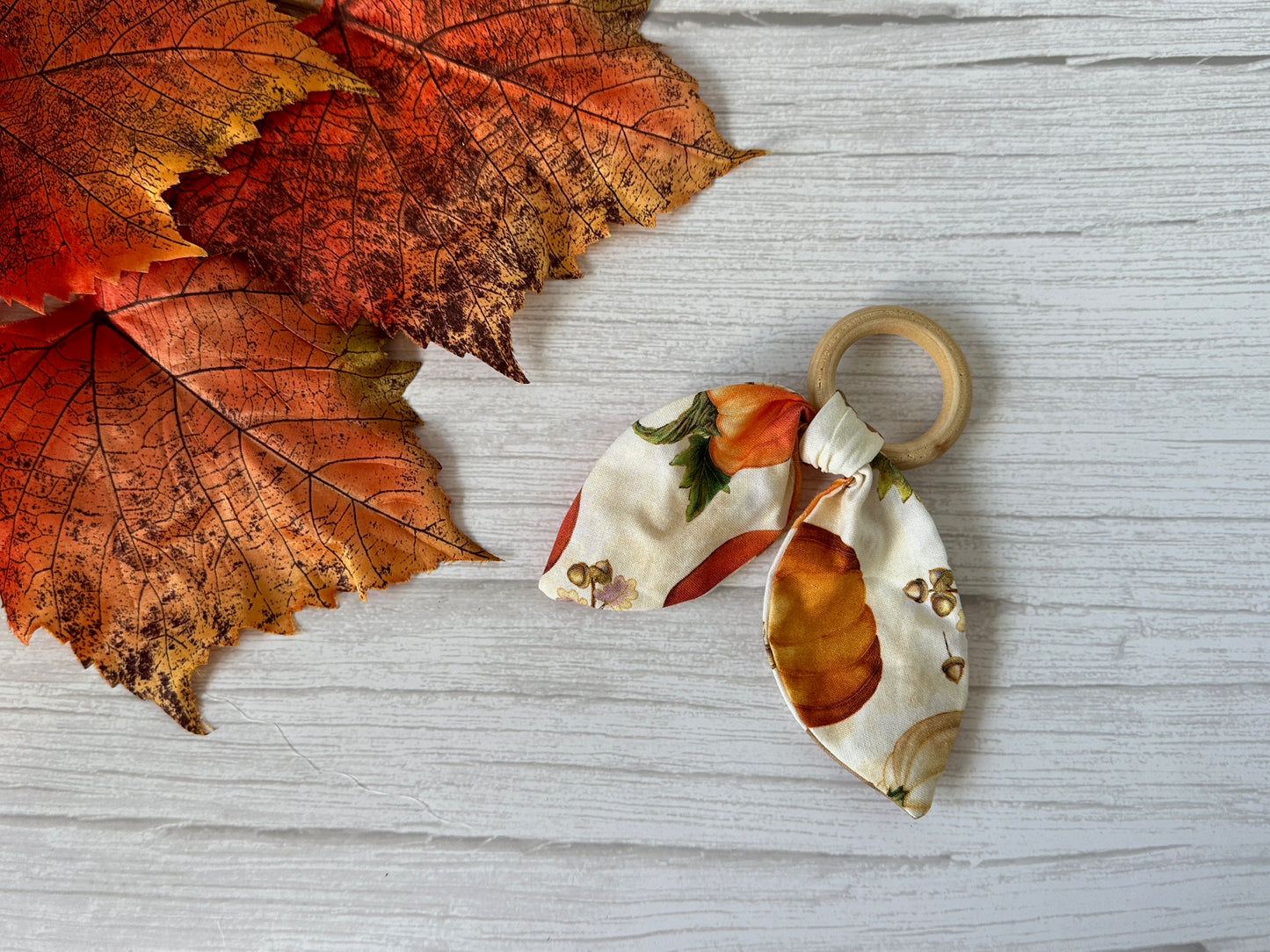 The Cotton & Bamboo Baby Teething Rattle - Autumn Pumpkins by Crafts by Kate features a handcrafted bamboo ring with a fabric bow, shown on a white wooden surface next to orange and red fall leaves.