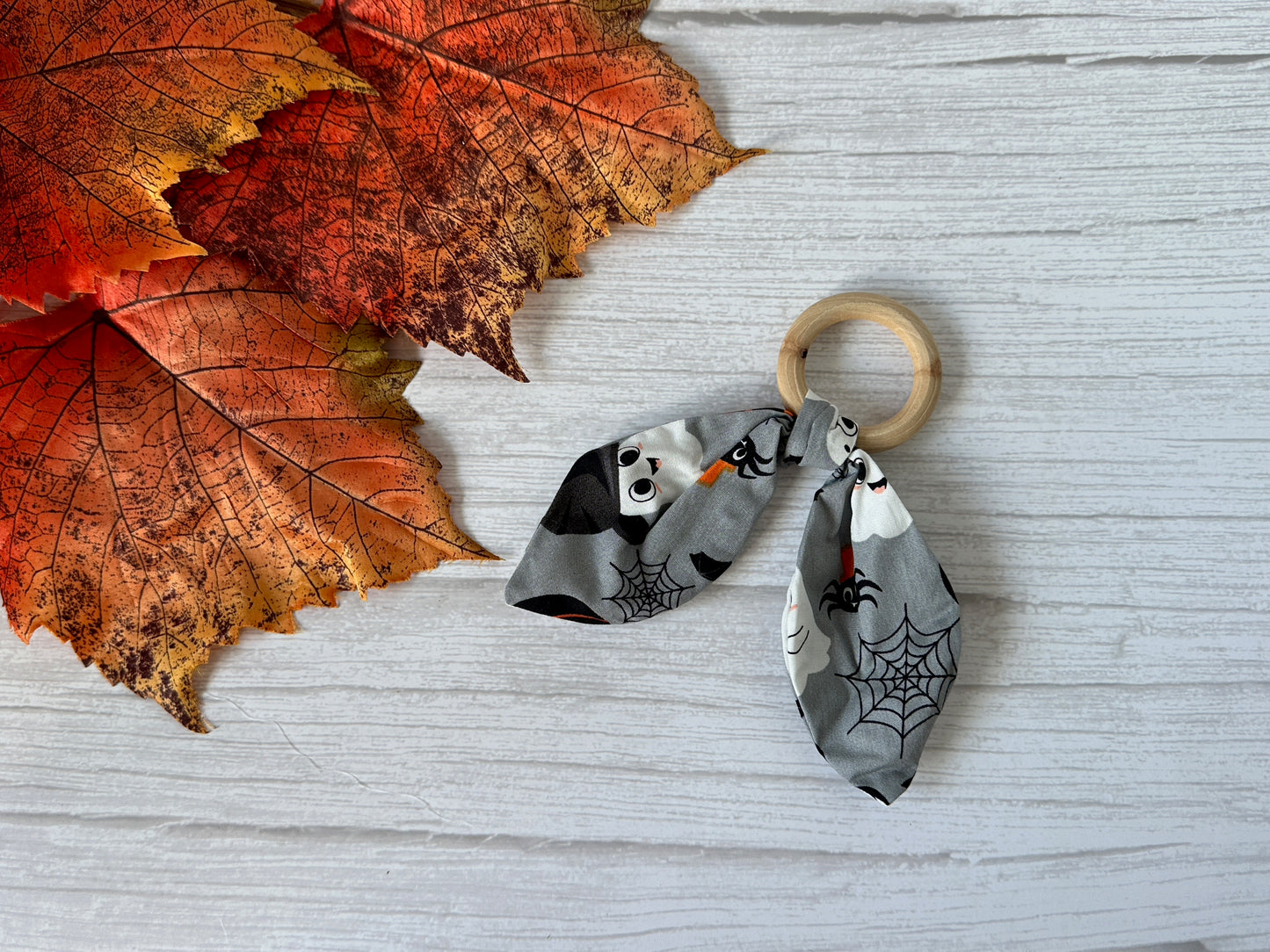 The Crafts by Kate Cotton & Bamboo Baby Teething Rattle - Grey Halloween, featuring ghosts, spiderwebs, and bats, rests beside orange autumn leaves on a white wooden surface.