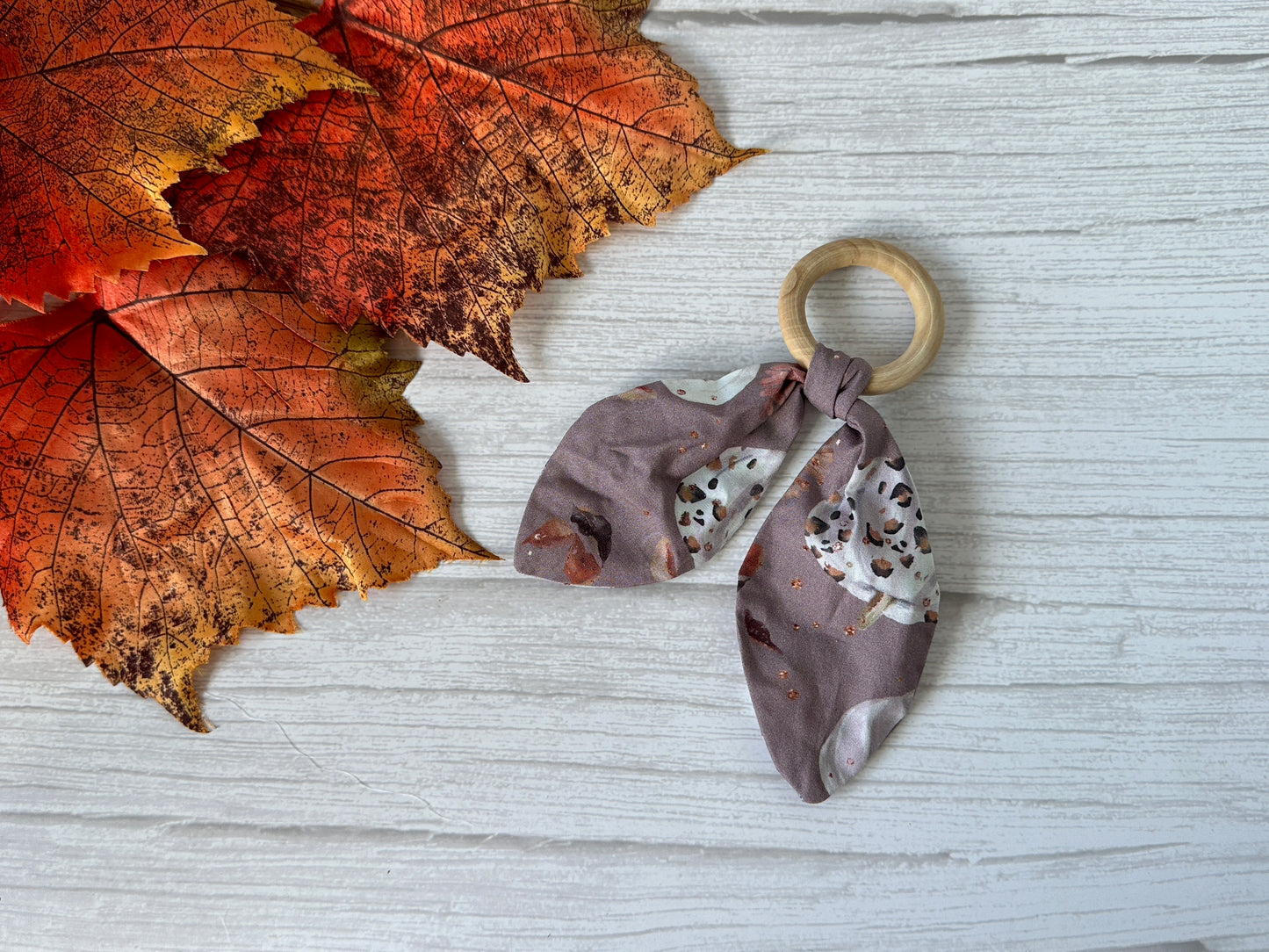 The Cotton & Bamboo Baby Teething Rattle - Leopard Print Pumpkins by Crafts by Kate rests on a white surface beside orange-red autumn leaves, featuring a wooden bamboo ring and knotted gray fabric with animal-themed prints.
