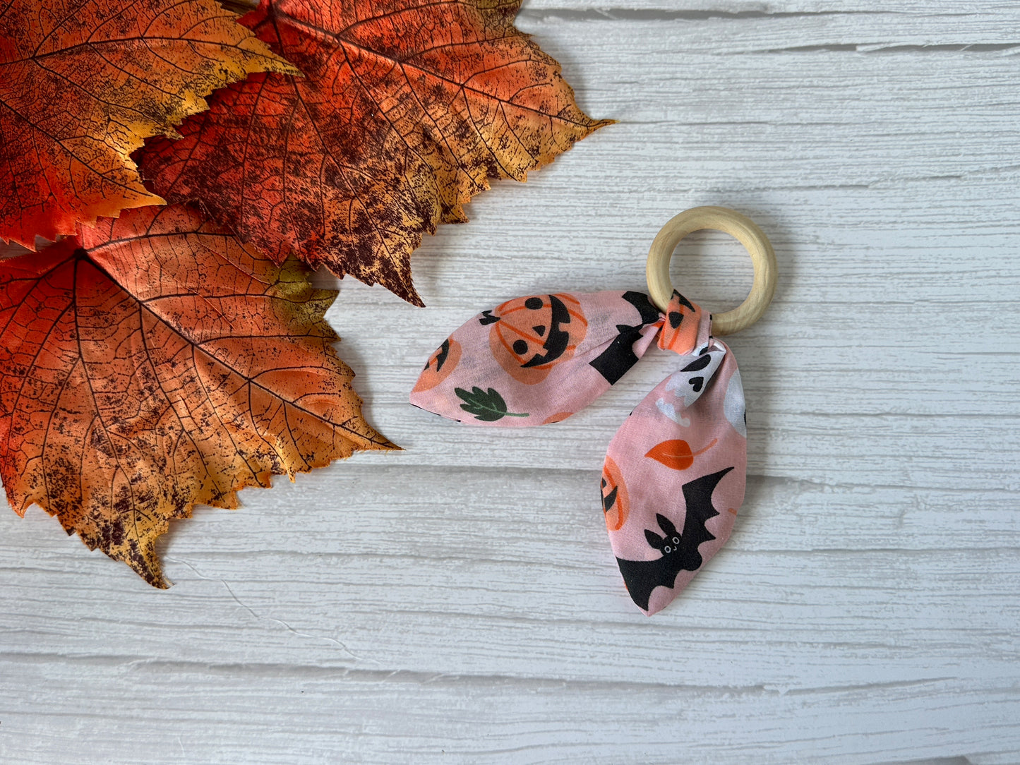 The Crafts by Kate Cotton & Bamboo Baby Teething Rattle – Pink Halloween features pumpkins and bats on fabric wrapped around a bamboo ring, pictured on a white wood surface with orange autumn leaves.