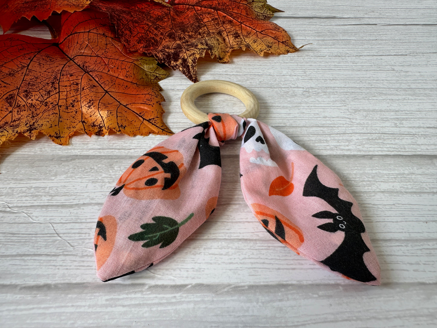 The Cotton & Bamboo Baby Teething Rattle - Pink Halloween by Crafts by Kate features a handcrafted bamboo ring with pink bunny ear fabric, decorated with pumpkins and bats, set against orange autumn leaves on a white wooden surface.