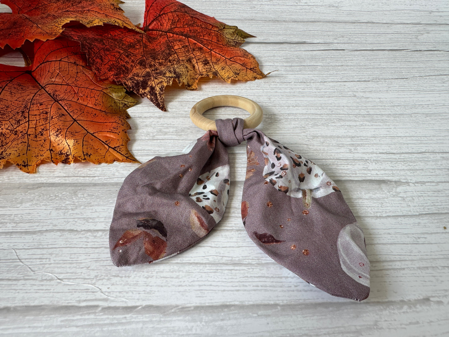 The Cotton & Bamboo Baby Teething Rattle - Leopard Print Pumpkins from Crafts by Kate rests on a white wooden surface, accented with autumn leaves. This handcrafted rattle features leopard print pumpkins on mauve fabric.