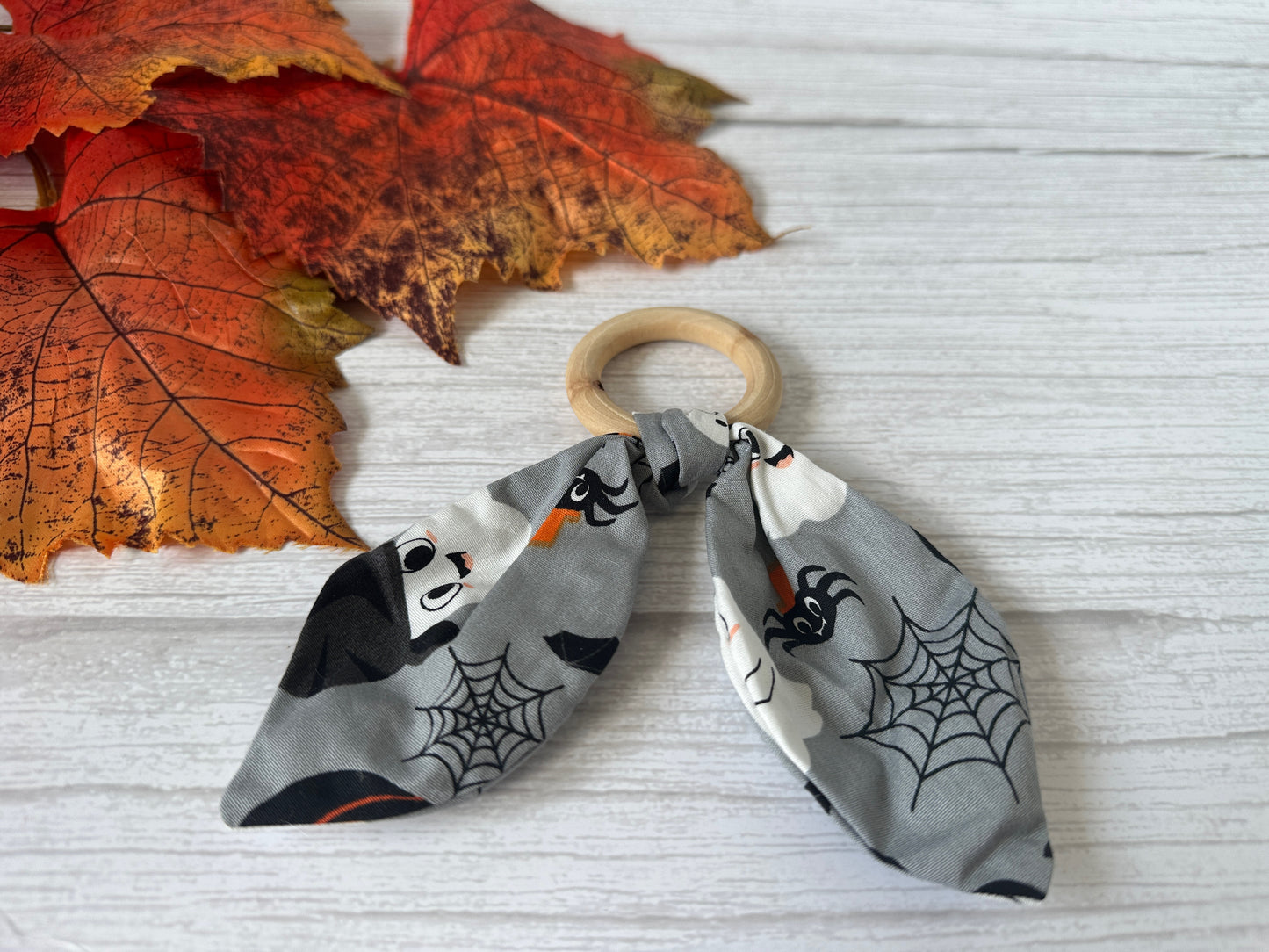 The Crafts by Kate Cotton & Bamboo Baby Teething Rattle - Grey Halloween, featuring spooky ghost, bat, and spider web fabric, rests on a white wooden surface beside orange autumn leaves.