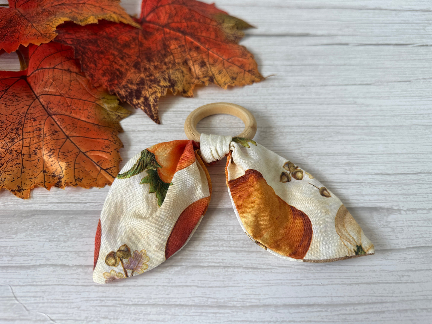The Cotton & Bamboo Baby Teething Rattle - Autumn Pumpkins by Crafts by Kate, featuring a handcrafted bamboo ring with a fabric bow in pumpkin and leaf prints, is displayed on a white wooden surface next to orange faux fall leaves.